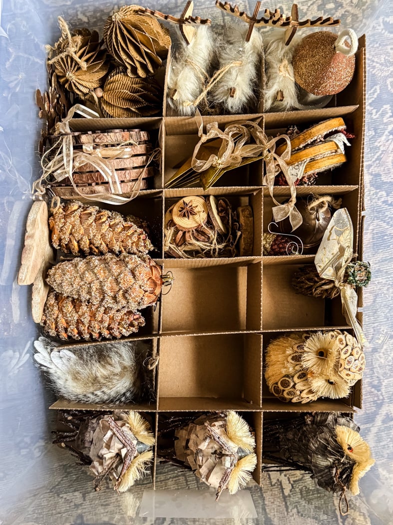 a cardboard box with dividers holds assorted rustic christmas ornaments, including pinecones, faux animal figures, dried oranges, stars, and wood slices, neatly organized in each compartment.