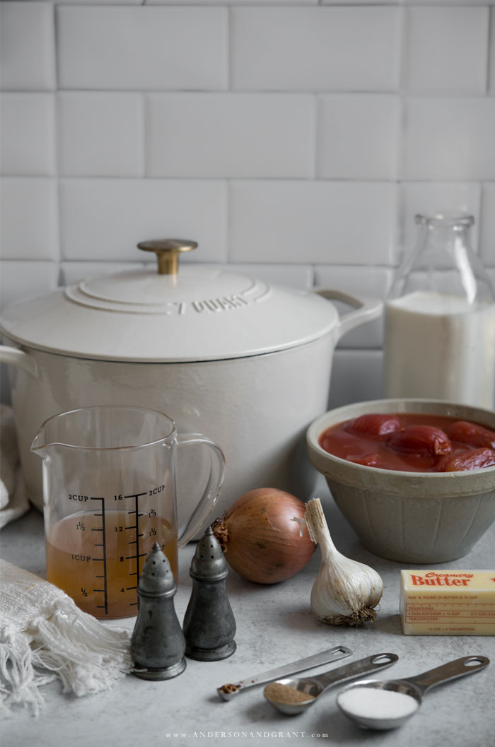 a white dutch oven sits on a counter with a bowl of whole tomatoes, a glass of broth, an onion, garlic, butter, salt, pepper shakers, a measuring spoon, and a bottle of milk in front of a white tile backsplash.