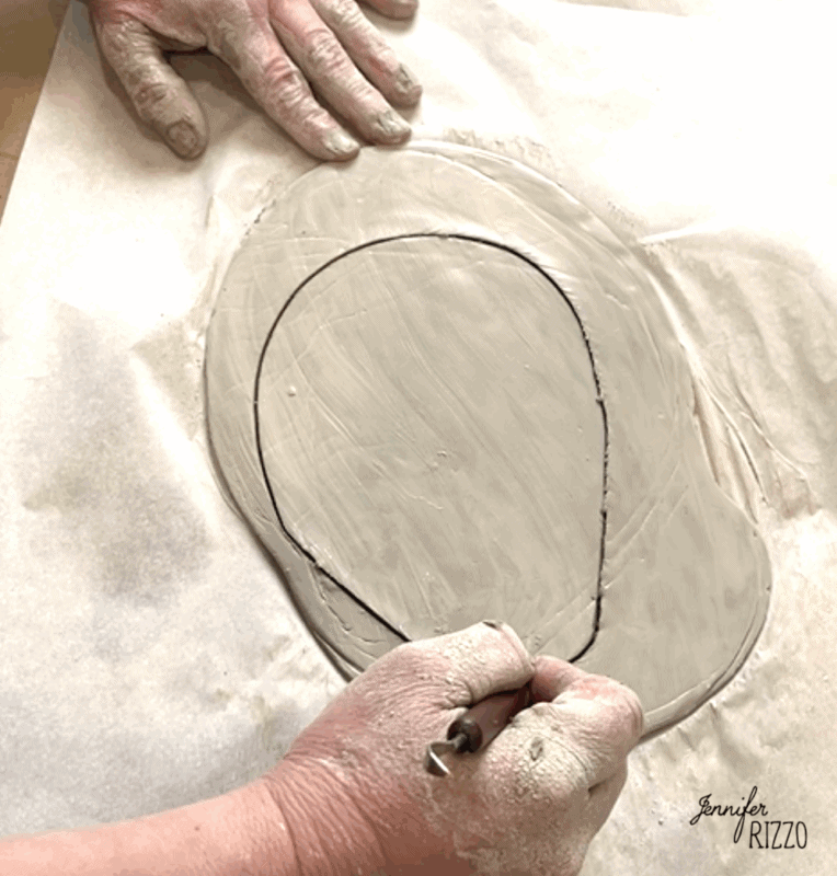 air dry clay strawberry trinket dish 3 a person uses a sculpting tool to trace an oval shape onto a slab of wet clay placed on parchment paper.