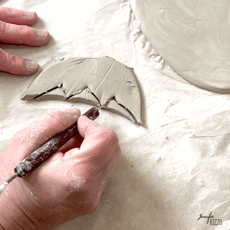 air dry clay strawberry trinket dish 5 a person uses a pointed tool to carve an umbrella shape from a sheet of gray clay on parchment paper.
