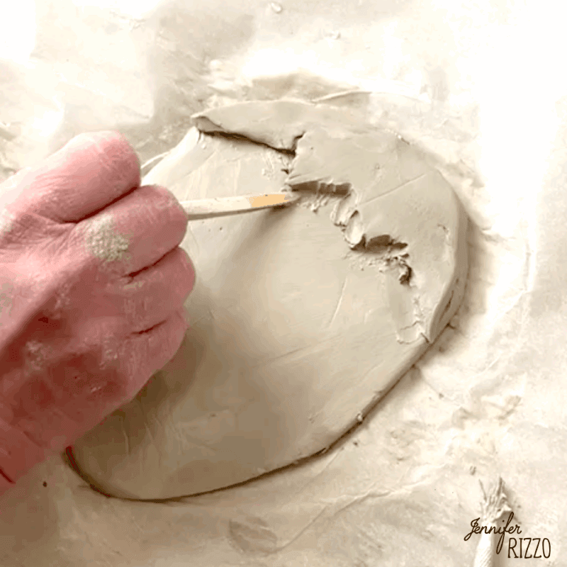 air dry clay strawberry trinket dish 7 a hand uses a wooden stick to carve into a flat slab of gray clay placed on a piece of parchment paper.