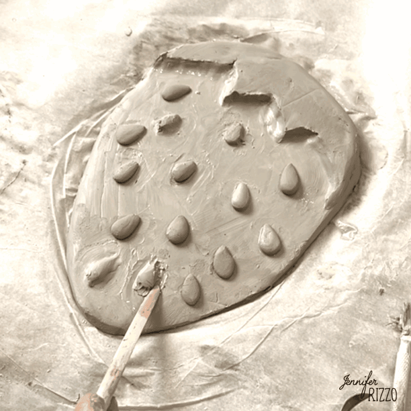 air dry clay strawberry trinket dish 8 a clay strawberry shape with added clay "seeds" is being sculpted on wax paper using a wooden stick.