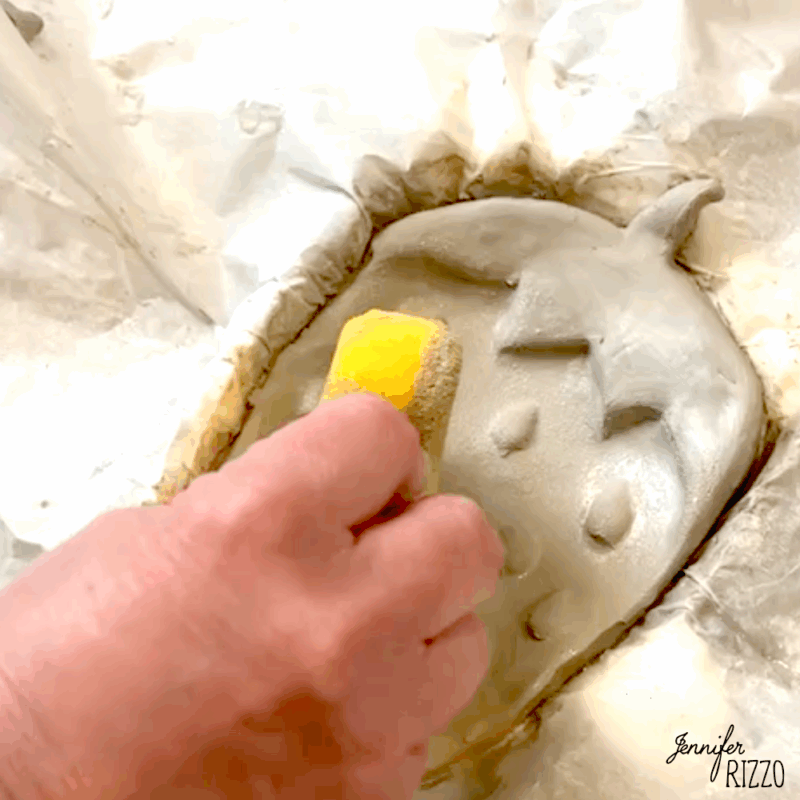 air dry clay strawberry trinket dish 9 a hand smooths the surface of a clay pumpkin shape with a yellow sponge; the clay rests on wrinkled parchment paper.