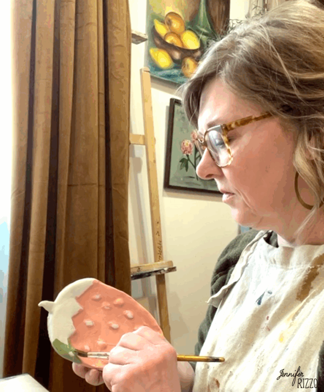 air dry clay strawberry trinket dish 11 a woman wearing glasses and an apron paints a ceramic piece shaped like a strawberry at a table in an art studio.