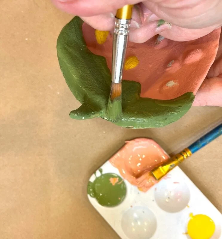 air dry clay strawberry trinket dish 12 a hand uses a paintbrush to apply green paint to a clay piece, with a paint palette featuring green, pink, and yellow paint visible nearby.