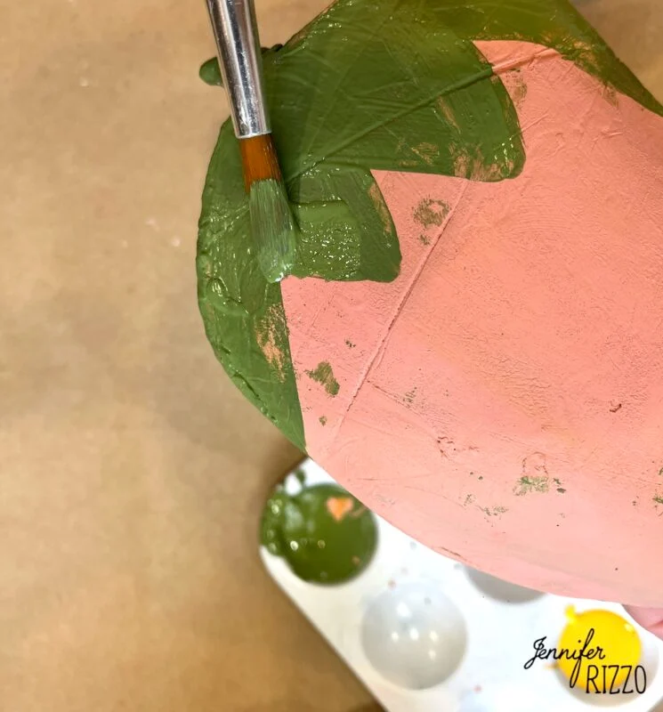air dry clay strawberry trinket dish 13 a hand uses a paintbrush to apply green paint to the top of a pink terracotta pot, with a paint palette visible nearby.