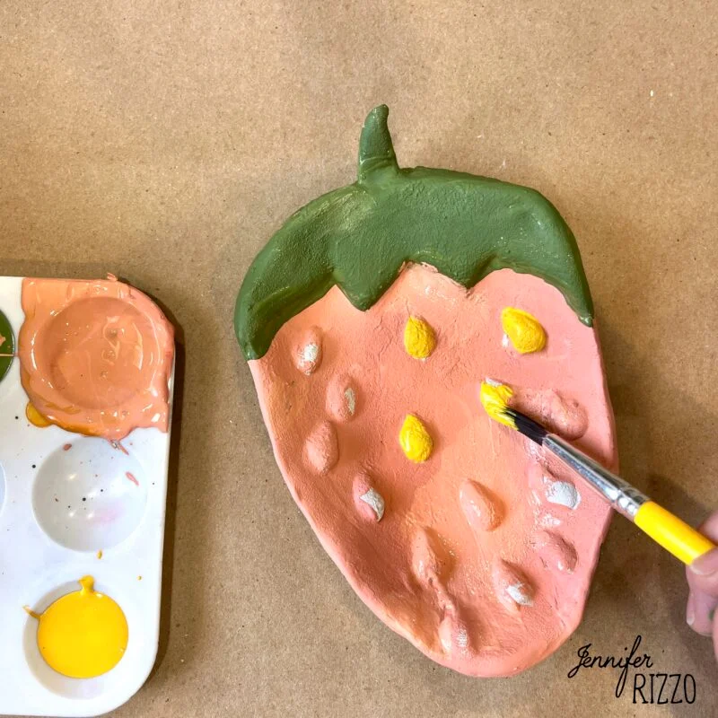 air dry clay strawberry trinket dish 14 a hand uses a paintbrush to paint yellow spots on a strawberry-shaped clay dish; a palette with pink and yellow paint is beside it.