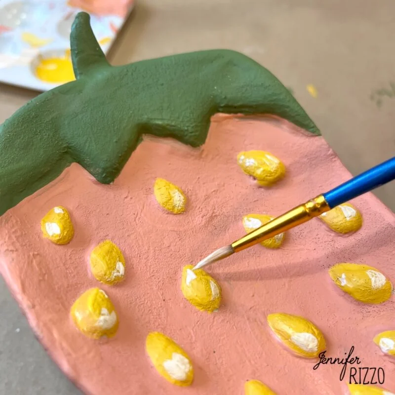 air dry clay strawberry trinket dish 15 a hand uses a small paintbrush to add white details to yellow seeds on a strawberry-shaped clay object painted pink and green.