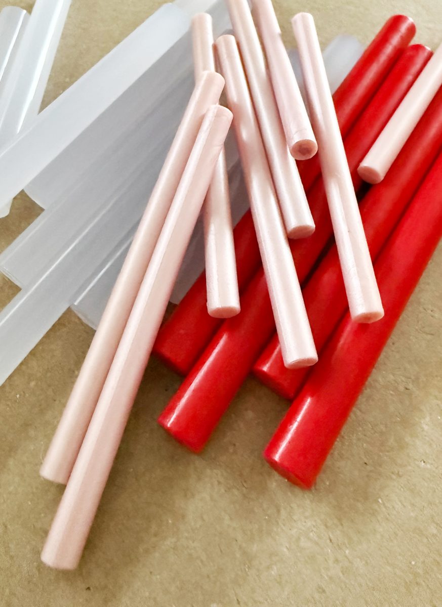 several glue sticks in three colors—clear, pale pink, and red—are spread out on a light brown surface, overlapping slightly. the sticks have a smooth, cylindrical shape.