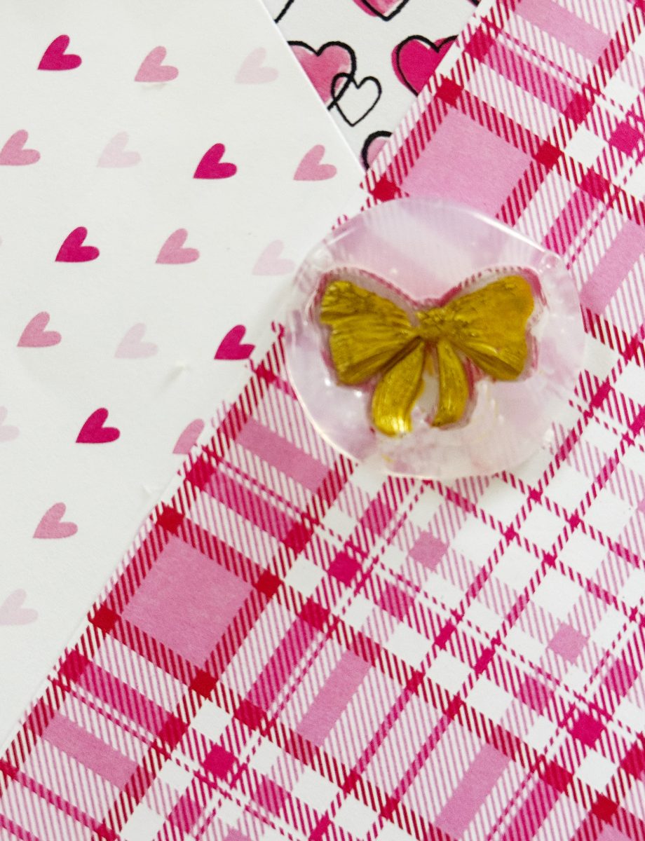 a gold bow sits on pink plaid fabric, next to white paper with pink hearts and another paper with black and pink outlined hearts.