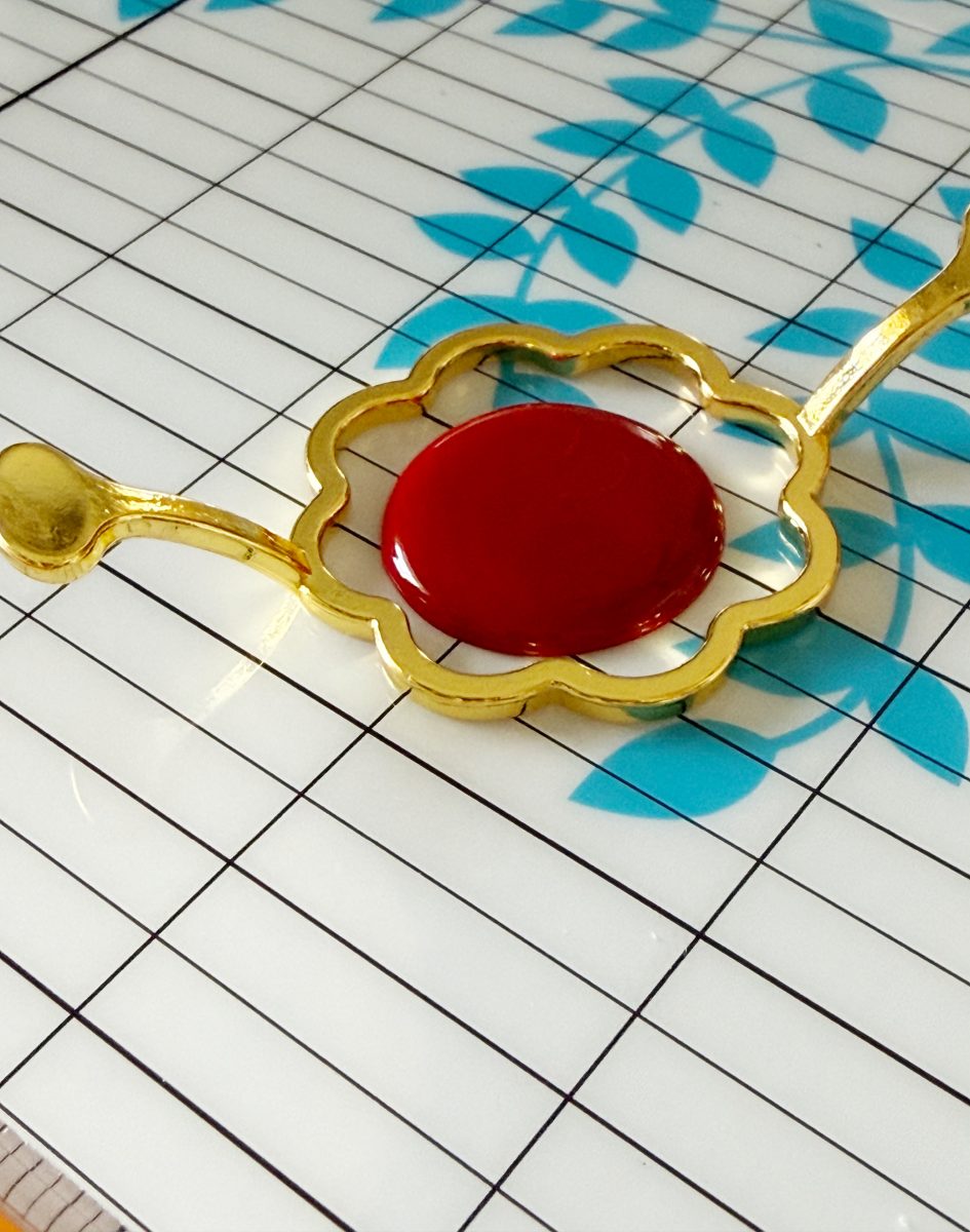 a gold flower-shaped frame holds a glossy red circle, resting on a white surface with black grid lines and blue leaf patterns.