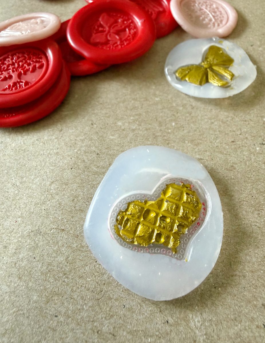 a translucent silicone stamp with a gold textured heart design rests on a brown surface, with red and pink wax seals and another stamp featuring a gold butterfly nearby.