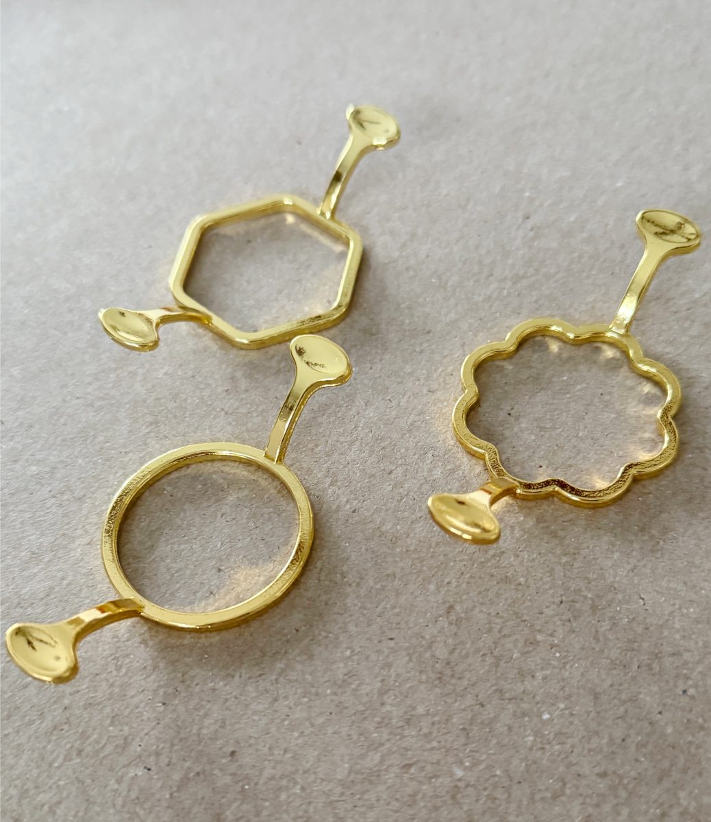 three gold-colored metal rings with handles, each in a different shape: hexagon, scalloped circle, and plain circle, lying on a neutral, textured surface.