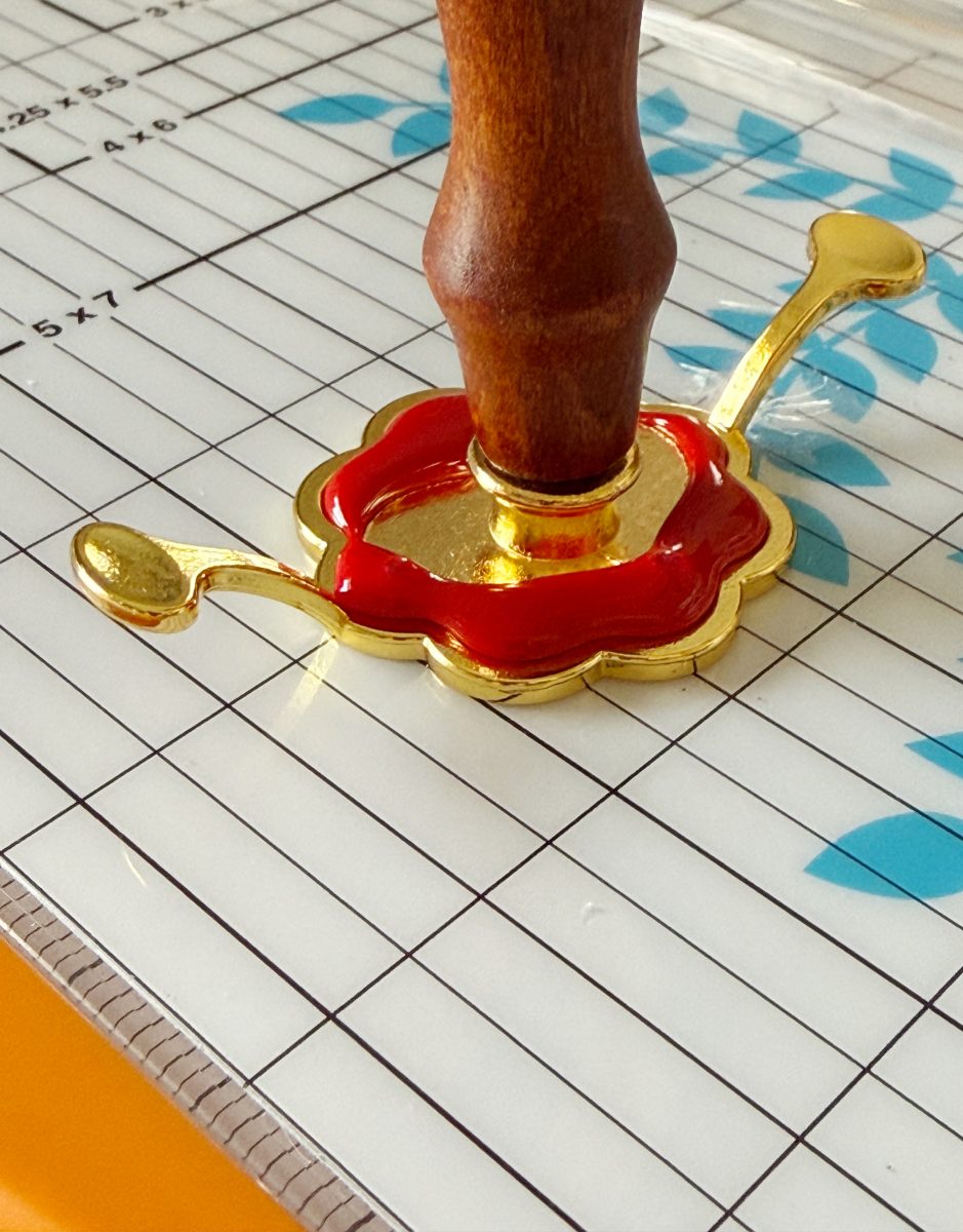 a wooden-handled wax seal stamp presses into bright red wax with gold embellishments on a patterned cutting mat featuring blue leaves and measurement lines.