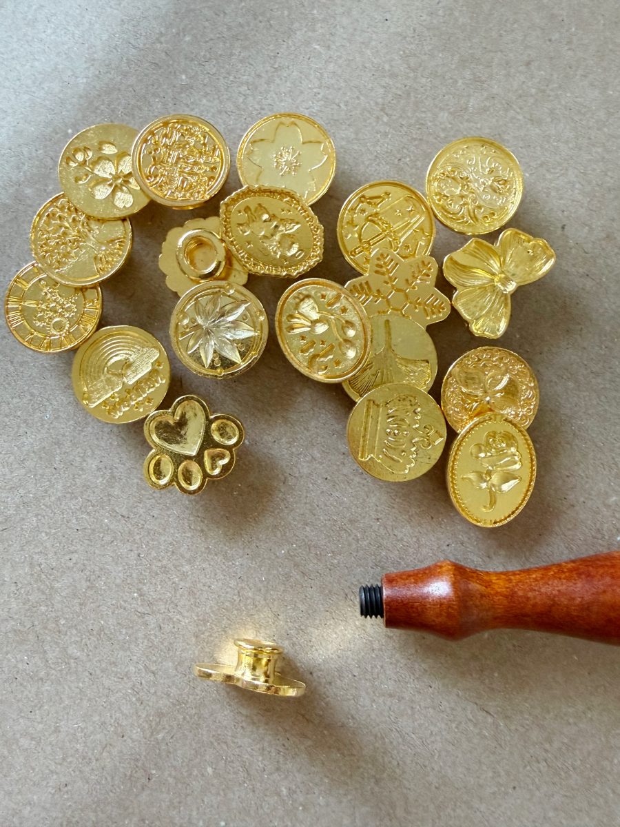 a collection of gold wax seal stamps with various floral and decorative designs is displayed on a brown surface. a wooden-handled stamp tool lies nearby with a seal attached.