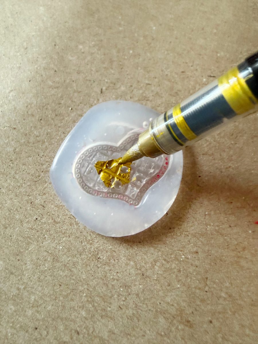 a pen with gold ink colors a small leaf design inside a translucent, heart-shaped silicone mold placed on a beige surface.