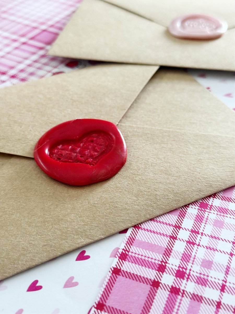 two brown envelopes are sealed with wax; one has a red heart-shaped seal and the other a pink seal. they are placed on pink and white patterned paper with small heart designs.