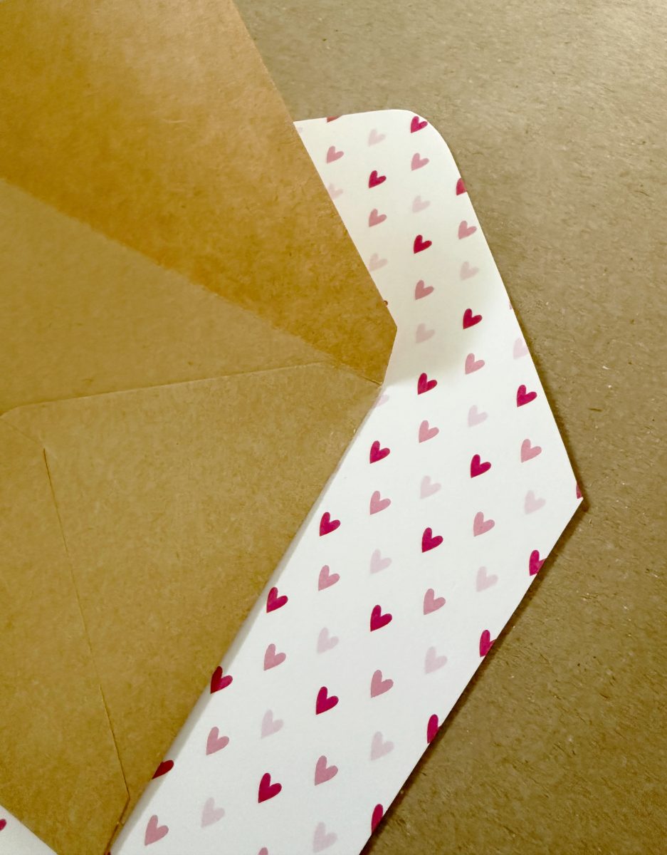 an open kraft paper envelope reveals its inner lining, which features a pattern of pink and red hearts on a white background. the envelope rests on a brown surface.