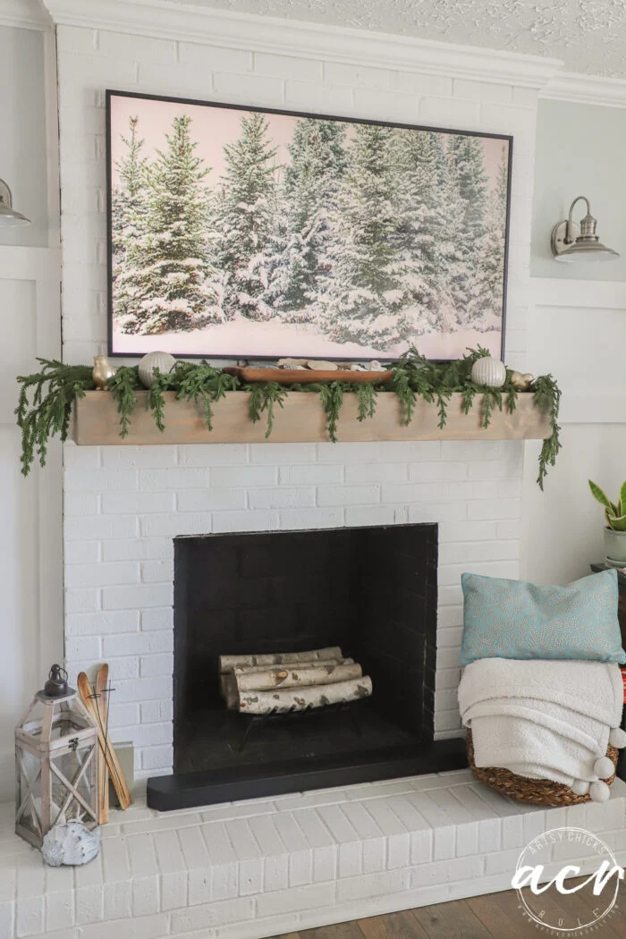 frame tv with green trees and snow on it with greenery on mantel