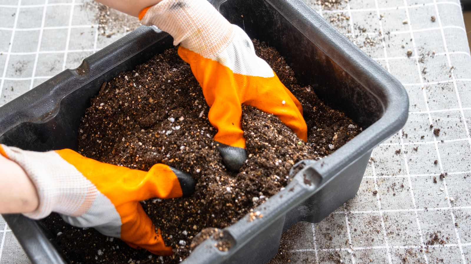 a close-up and overhead shot of a person's hands wearing orange gloves, in the process of mixing a high quality potting mix