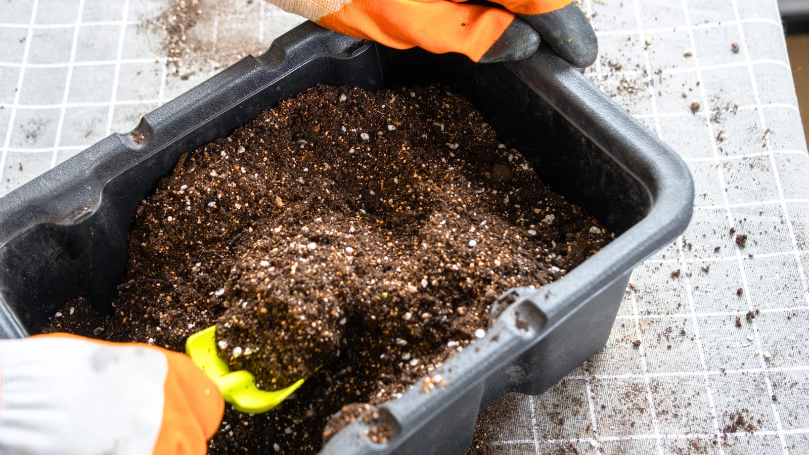 a gardener wearing orange gloves mixes potting soil in a large black bucket using a green trowel to transplant houseplants.