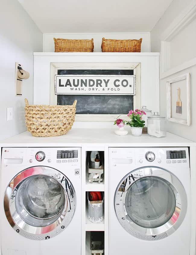 how i wake up my house 9 a modern laundry room with two front-load washing machines under a counter displaying a decorative sign reading "laundry co. wash, dry, & fold," a wicker basket, small potted flowers,