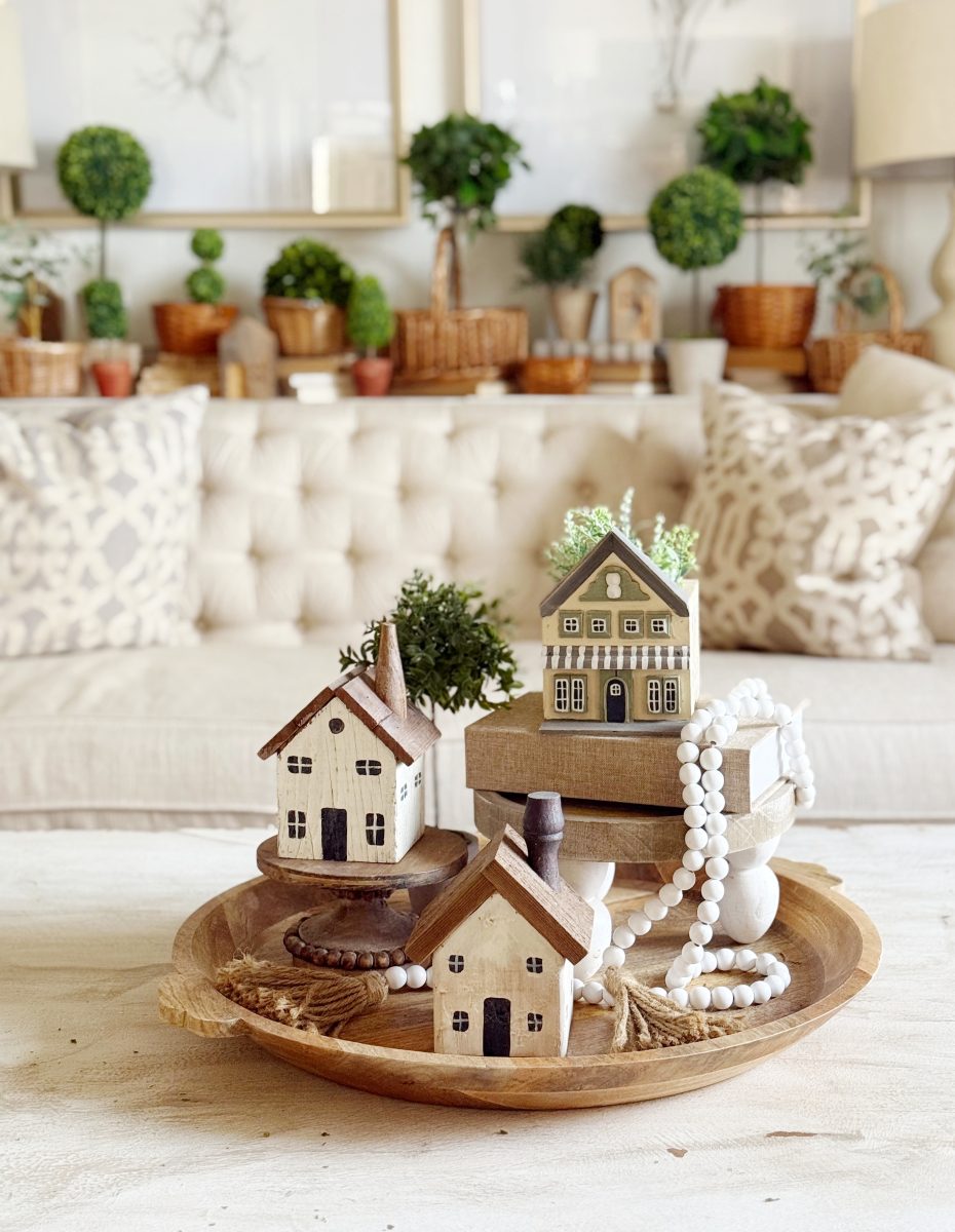 how i wake up my house 14 a wooden tray with small decorative house figurines, books, greenery, and beads sits on a coffee table in a cozy living room with a beige sofa and potted plants in the background.