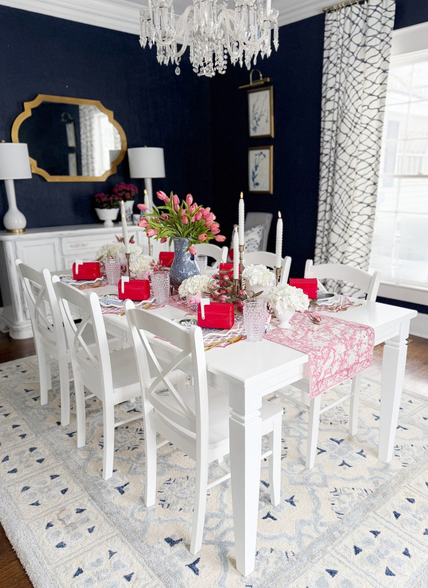 a dining room with dark blue walls and white trim features a white table set for six with red napkins, pink flowers, elegant glassware, a pink table runner, and a crystal chandelier above. large windows let in natural light.