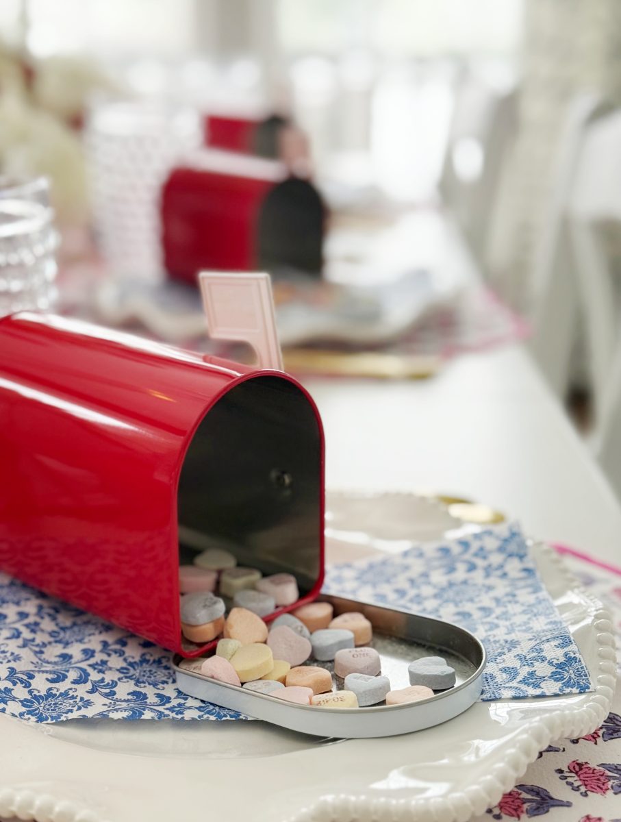 a small red mailbox lies open on its side, spilling colorful heart-shaped candies onto a blue and white patterned napkin on a white table set for a meal.