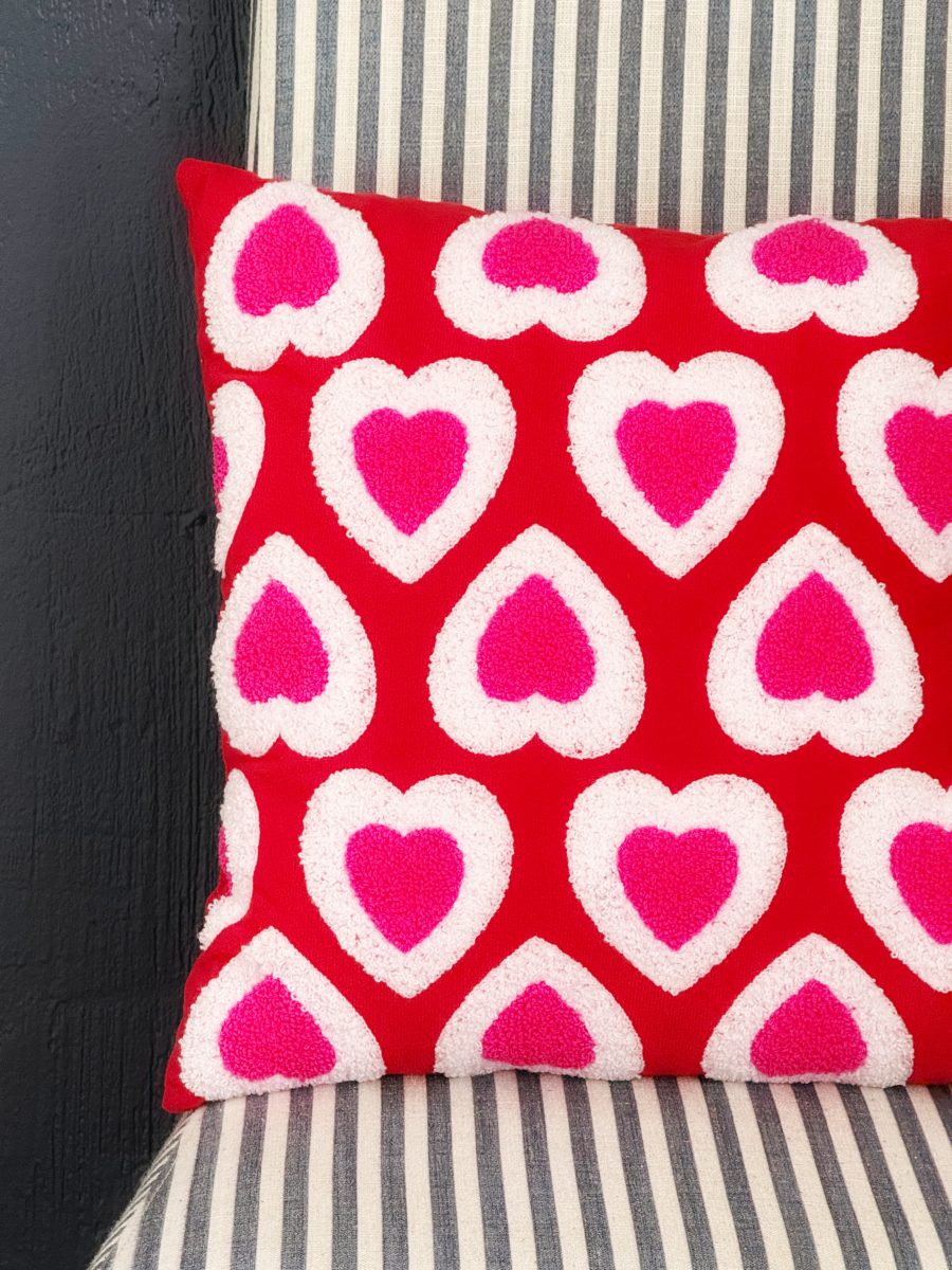 a red pillow with white and pink heart patterns rests on a chair with a black and white striped cushion, next to a dark textured wall.