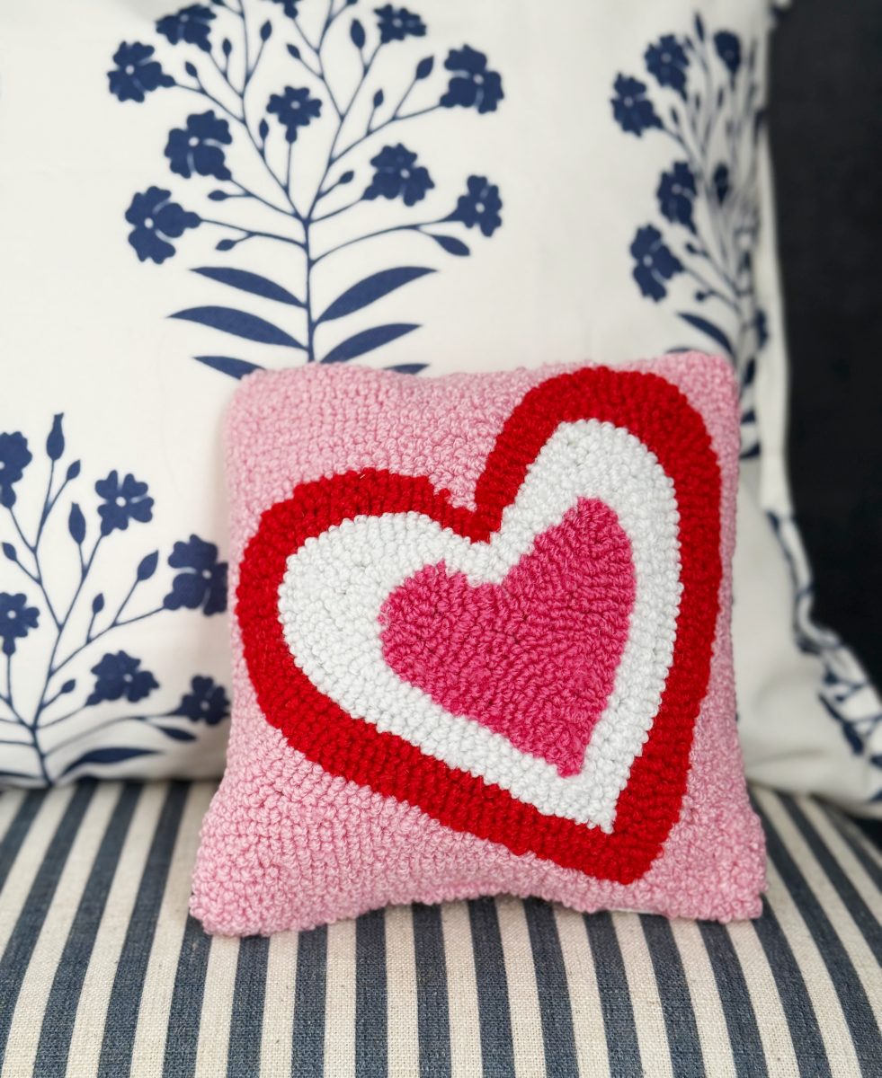 a small square pillow with a pink, red, and white heart design sits on a striped cushion, with a white pillow featuring blue floral patterns in the background.