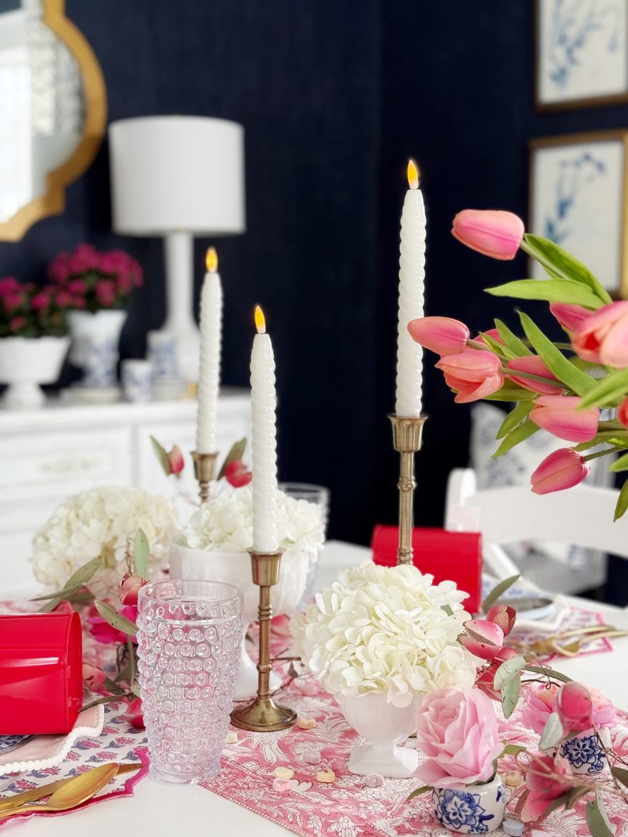 a table set with pink and white floral arrangements, bubble glasses, brass candlesticks holding lit white candles, and pink tulips, with a dark wall and white furniture in the background.