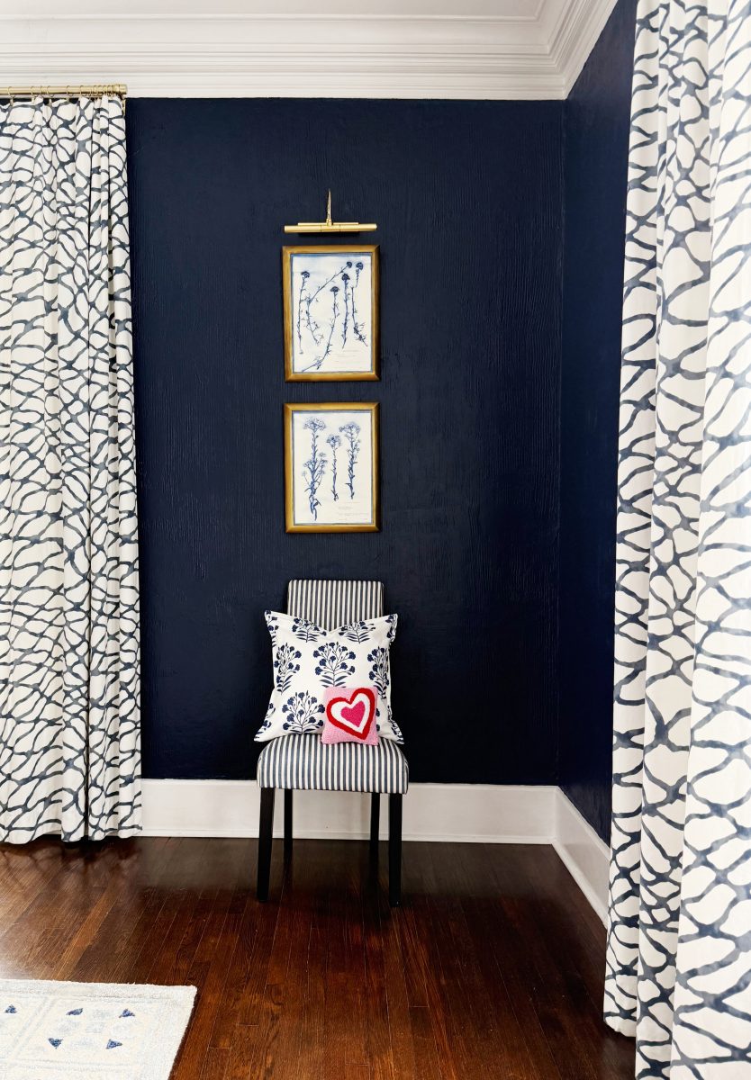 a corner with dark blue textured walls, patterned curtains, and a black-striped chair topped with a floral pillow and a pillow featuring a red heart. two framed botanical prints hang above the chair. wood floor.