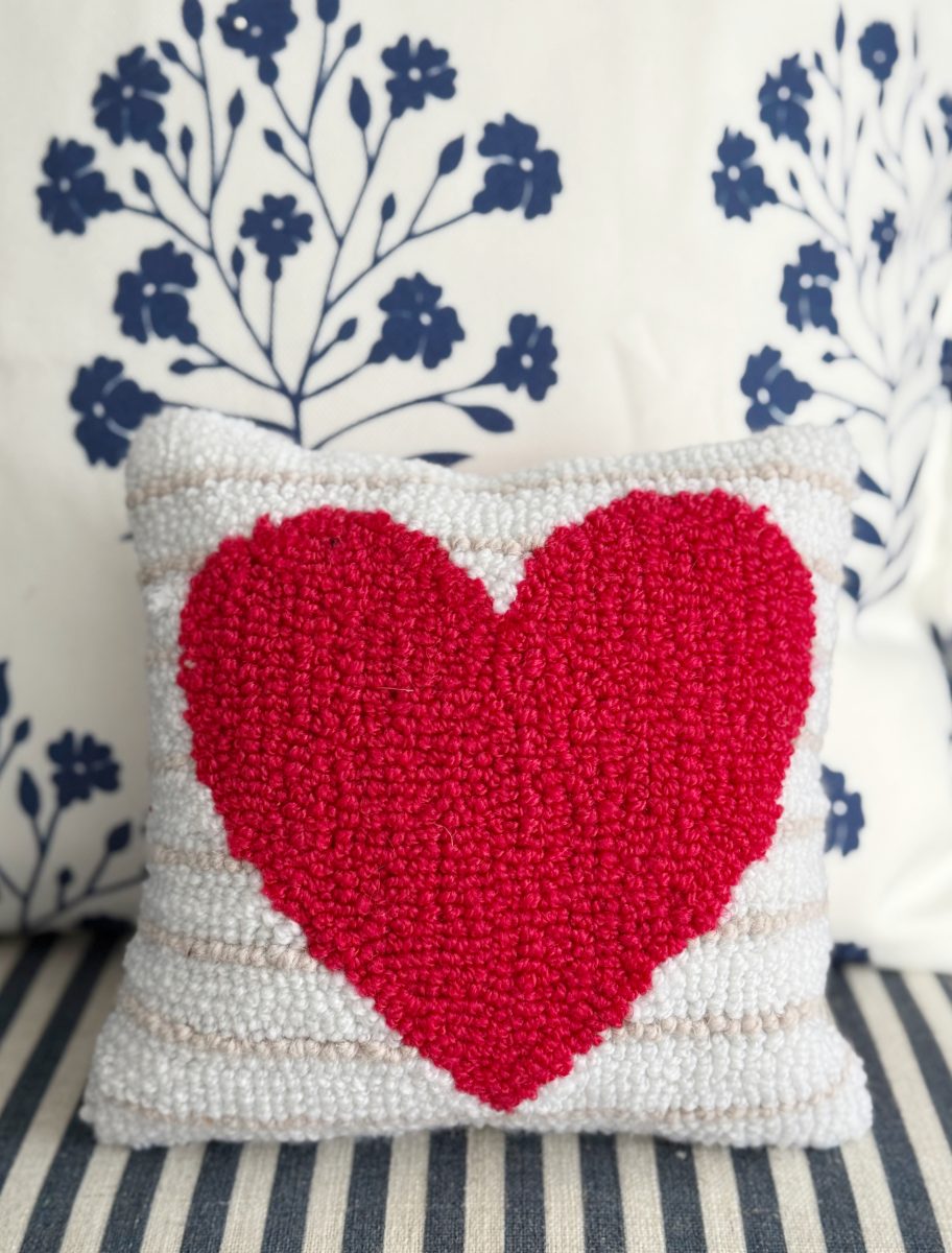 a square pillow with a large red heart design sits on a striped cushion. the pillow has a textured, handmade look, and behind it is a white cushion with a blue floral pattern.