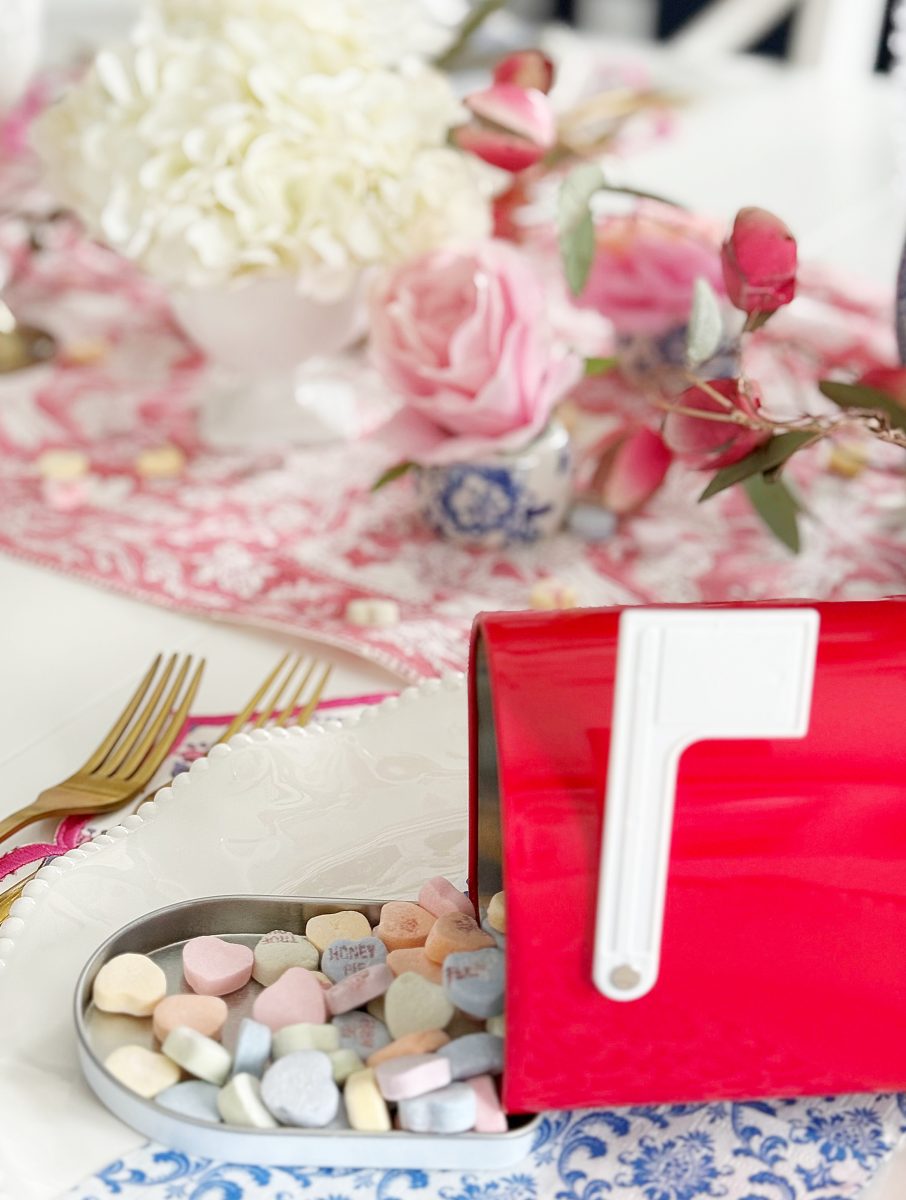 a small red mailbox tipped over, spilling pastel candy hearts onto a white plate. pink roses, white flowers, gold utensils, and a pink patterned tablecloth decorate the scene in the background.