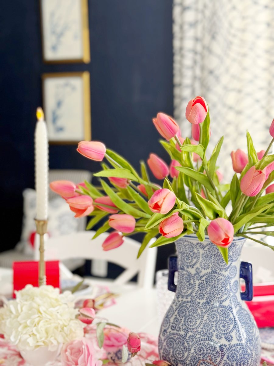 a blue and white patterned vase holds a bouquet of pink tulips on a table set with a white candle, white hydrangeas, and pink roses. the background features a dark wall and framed art.