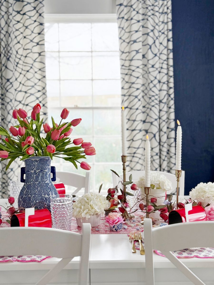 a dining table is decorated with pink tulips in a blue patterned vase, white flowers, glassware, pink and red party favors, gold candlesticks with white candles, and a patterned tablecloth. sheer curtains cover the window behind.