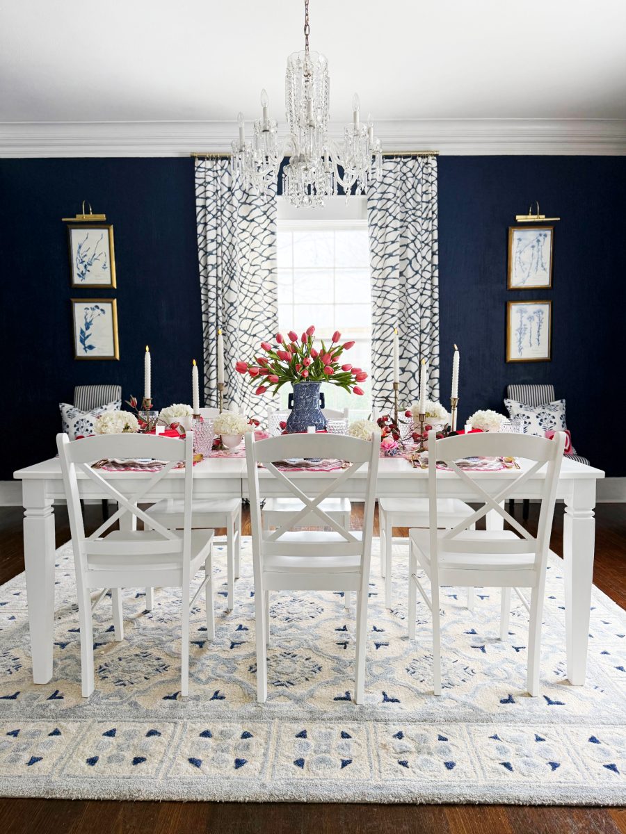 a dining room with a white table and six white chairs, set with flowers, candles, and tableware. a crystal chandelier hangs above, and the room features navy blue walls, patterned curtains, and framed artwork.