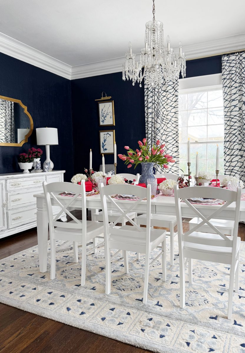 elegant dining room with dark blue walls, white trim, and a crystal chandelier. a white table and chairs sit on a patterned rug, decorated with pink flowers and candles. large windows have patterned curtains.