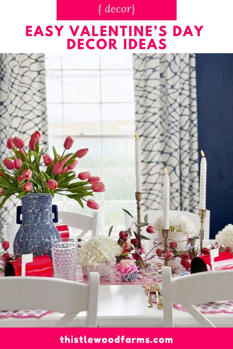 a dining table decorated for valentine’s day with pink tulips in a blue vase, white candles, red napkins, and white flowers. the background features patterned curtains and a bright window.