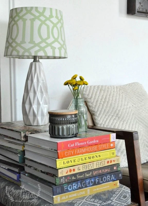 different ways to use vintage books in decorating 15 a white geometric table lamp, a small vase with yellow flowers, and a grey candle sit on a stack of books next to a textured pillow on a wooden chair.