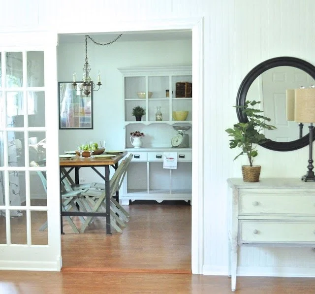ways to repurpose china cabinets 24 a dining area with a wooden table and chairs, a white hutch, and chandelier, viewed from an adjacent light and bright cottage living room makeover featuring a round mirror and a potted plant on the console table.