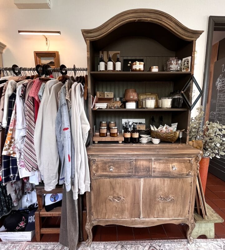 ways to repurpose china cabinets 28 a wooden hutch, showcasing candles and jars, stands beside a clothing rack with shirts, sweaters, and scarves—offering stylish inspiration and repurposing ideas for outdated furniture in a chic boutique setting.