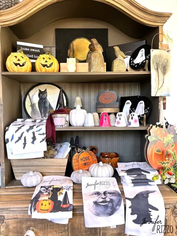 ways to repurpose china cabinets 29 a wooden shelf displays halloween-themed decorations, including pumpkins, ghost figurines, black cat art, and holiday towels, with a “jennifer rizzo” label on several items.