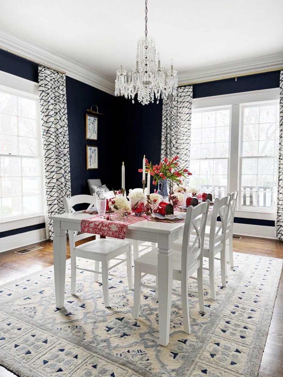 bright dining room with dark blue walls, large windows with patterned curtains, a crystal chandelier, white table and chairs, a patterned rug, and a table set with pink and red flowers, candles, and festive decor.