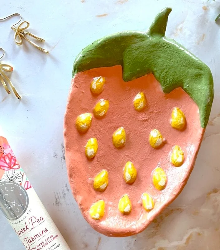air dry clay strawberry trinket dish 17 a ceramic dish shaped and painted like a strawberry with yellow seed details sits on a light surface next to a tube of lotion and a pair of gold earrings.