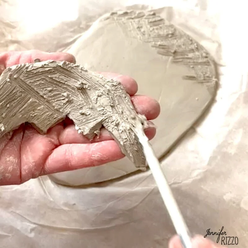 air dry clay strawberry trinket dish 6 a hand holds a piece of textured clay while another hand uses a tool to carve patterns into a larger clay slab on parchment paper.