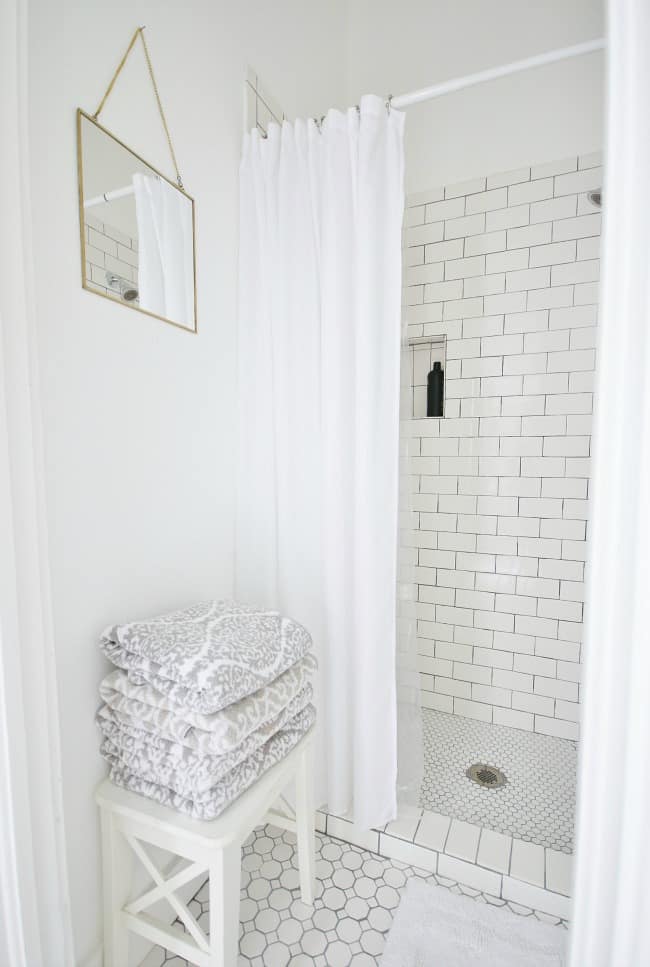 a clean, bright bathroom with white subway tiles on the wall, hexagonal tiles on the floor, a white shower curtain, and a stack of towels on a small stool next to a mirror.