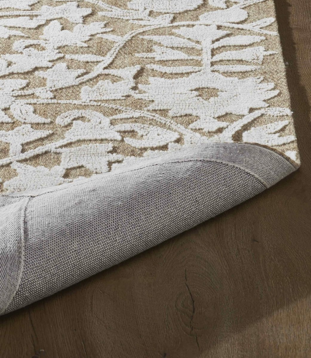 a close-up of a beige and white patterned rug with its corner slightly lifted, revealing a gray non-slip backing on a dark wooden floor—perfect for adding comfort beneath your spring mantel decor.