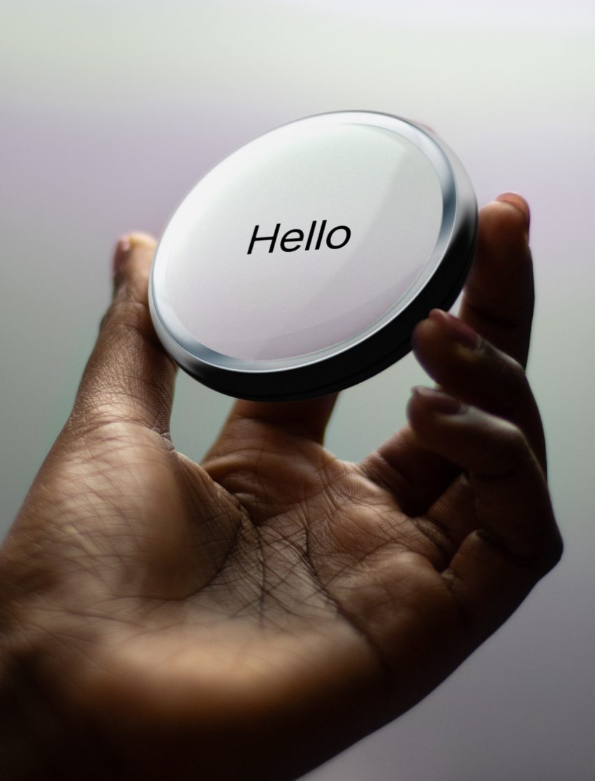 Image of a hand holding part of the Cognitive Bloom device in the form of a palm-sized smooth chrome disc with a white screen and the word hello in simple sans serif text